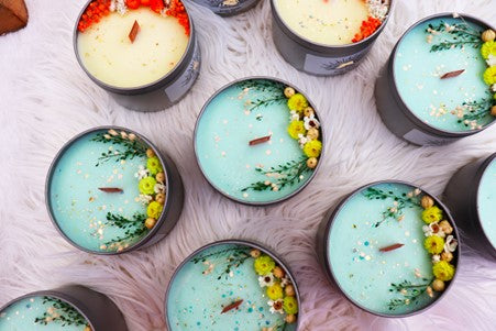 A picture of several homemade candles with botanicals and glitter in them lined up on a pink background