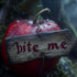 A close-up image of a red apple with a wooden tag attached to it, bearing the inscription 'bite me' in a rustic font, set against a blurred background with moss and foliage.