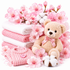 A styled gift set featuring a plush teddy bear wearing a pink bow, soft pink and white folded towels, a pink knit sweater, cotton bolls, and pink blossoms on a clean white background.