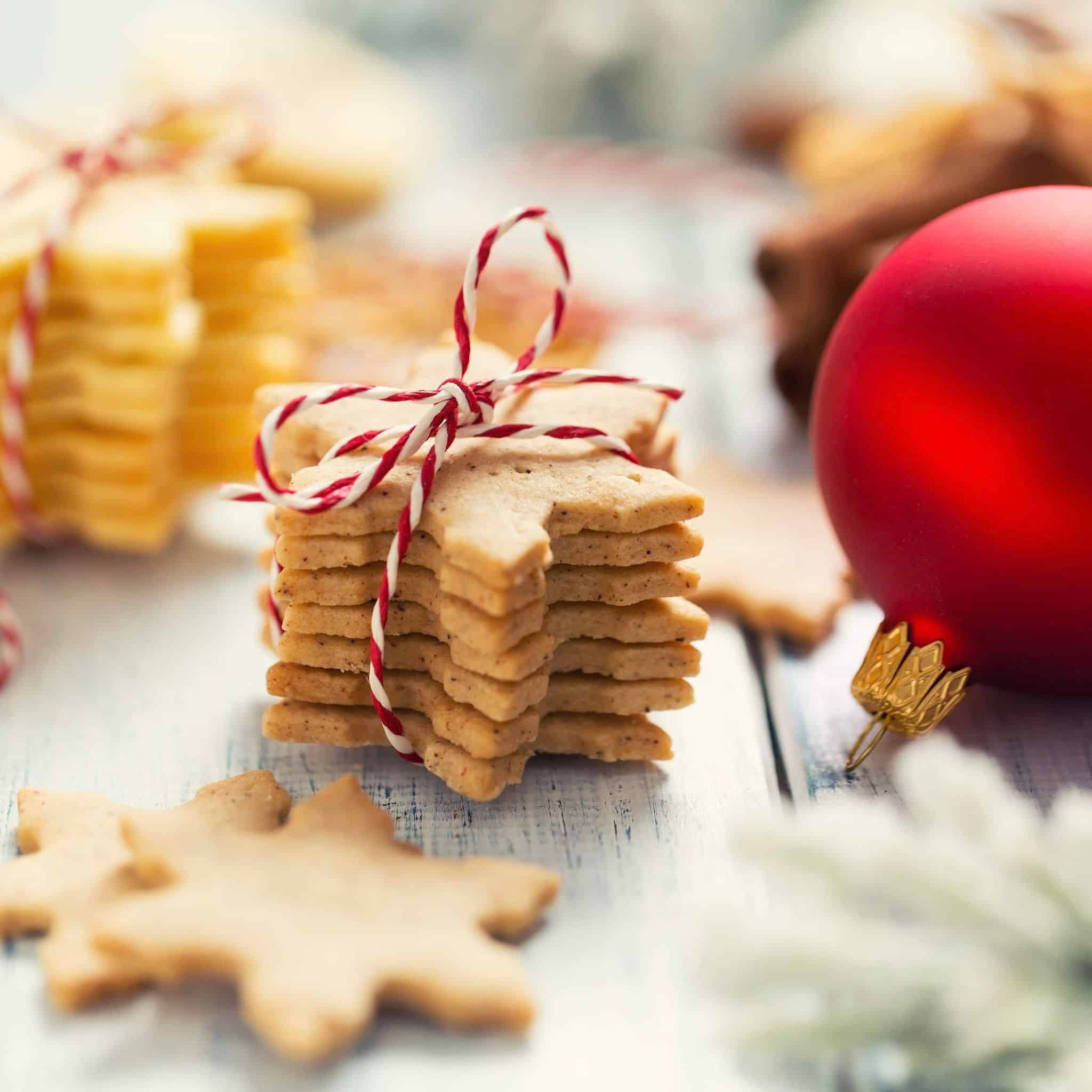 Stack of Christmas cookies tied with a red and white string, alongside a red ornament and more cookies in the background, on a surface with a blurred Christmas-themed backdrop.