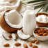 A glass bottle filled with white coconut milk stands on a light surface beside a halved coconut, sliced coconut pieces, almonds, and small bowls of coconut oil and almonds, with soft natural styling in the background.