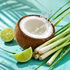 A halved coconut with white flesh and brown husk sits on a turquoise surface with water splashing from inside, surrounded by lime wedges, green stalks, and lemongrass.