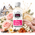 A bottle of Fizzy Whiz Cosy Cottage Fragrance Oil centered in a styled holiday scene with pink and cream roses, white berries, vanilla beans, amber pieces, a knit scarf, and a snowy house background.