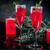Two fluted glasses filled with a red cranberry cocktail, garnished with a sprig of rosemary, alongside a red candle and decorative foliage on a dark background.