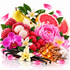 A vibrant, stylized arrangement of fragrance ingredients on a white reflective background, including pink lychee blossoms, red lychees, pink raspberries, a sliced pink grapefruit, a yellow pear, white orchids, vanilla beans, amber resin cubes, cinnamon sticks, and pink flower petals.