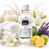 A bottle of Fizzy Whiz Fluffy Towels Fragrance Oil centered on a white background, surrounded by a lit candle, folded white towels, white lilies, purple lavender, a yellow lemon, and a green apple. The bottle has a white cap and a white label with black and pink branding.