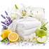 A styled spa-like arrangement with a stack of folded white towels, a white lily flower, purple lavender sprigs, a green apple, a lemon half, white flowers, clear faceted gems, and a lit candle in a glass jar on a white background.