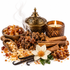A styled still life of natural fragrance ingredients on a white background, including amber resin pieces, cinnamon sticks, vanilla pods, a white flower, incense, and an ornate brass incense burner with a lit candle nearby.