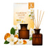 Garden Glow Amber & Jasmine reed diffuser set, featuring an amber glass bottle with natural wood reeds, its cream and orange packaging box, and decorative white jasmine flowers and amber crystals on a white background.