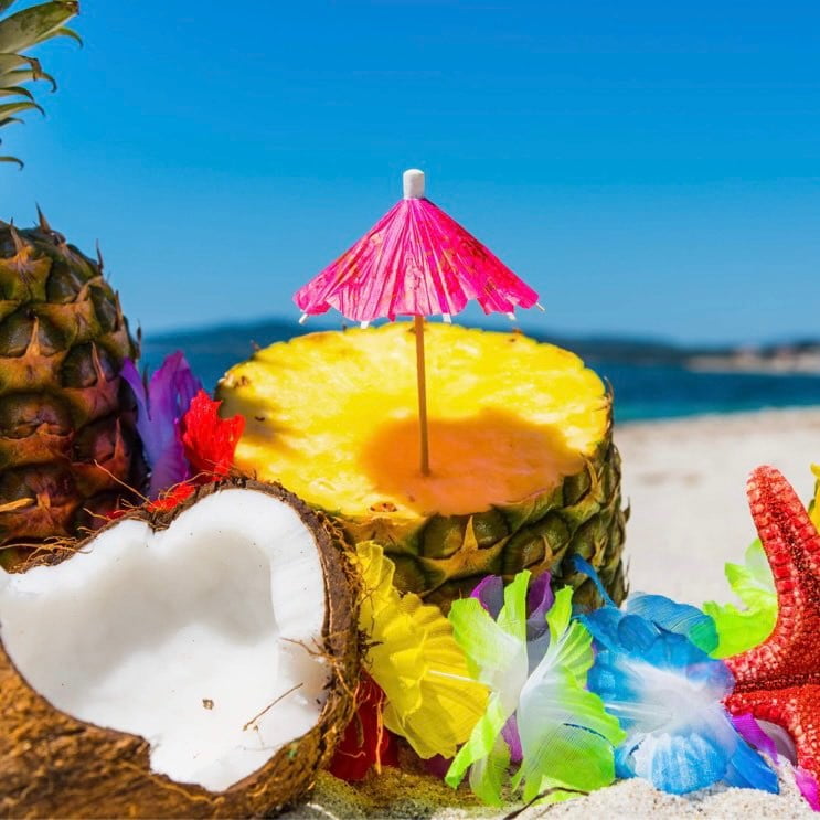 A tropical-themed arrangement featuring a sliced coconut, a whole pineapple, and various colorful flowers on a sandy beach backdrop. A small pink umbrella is placed on top of the pineapple slice.