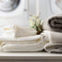 A stack of folded laundry including white and grey towels with a small bouquet of flowers on top, all placed on a surface with a reed diffuser in the background emitting fragrance.