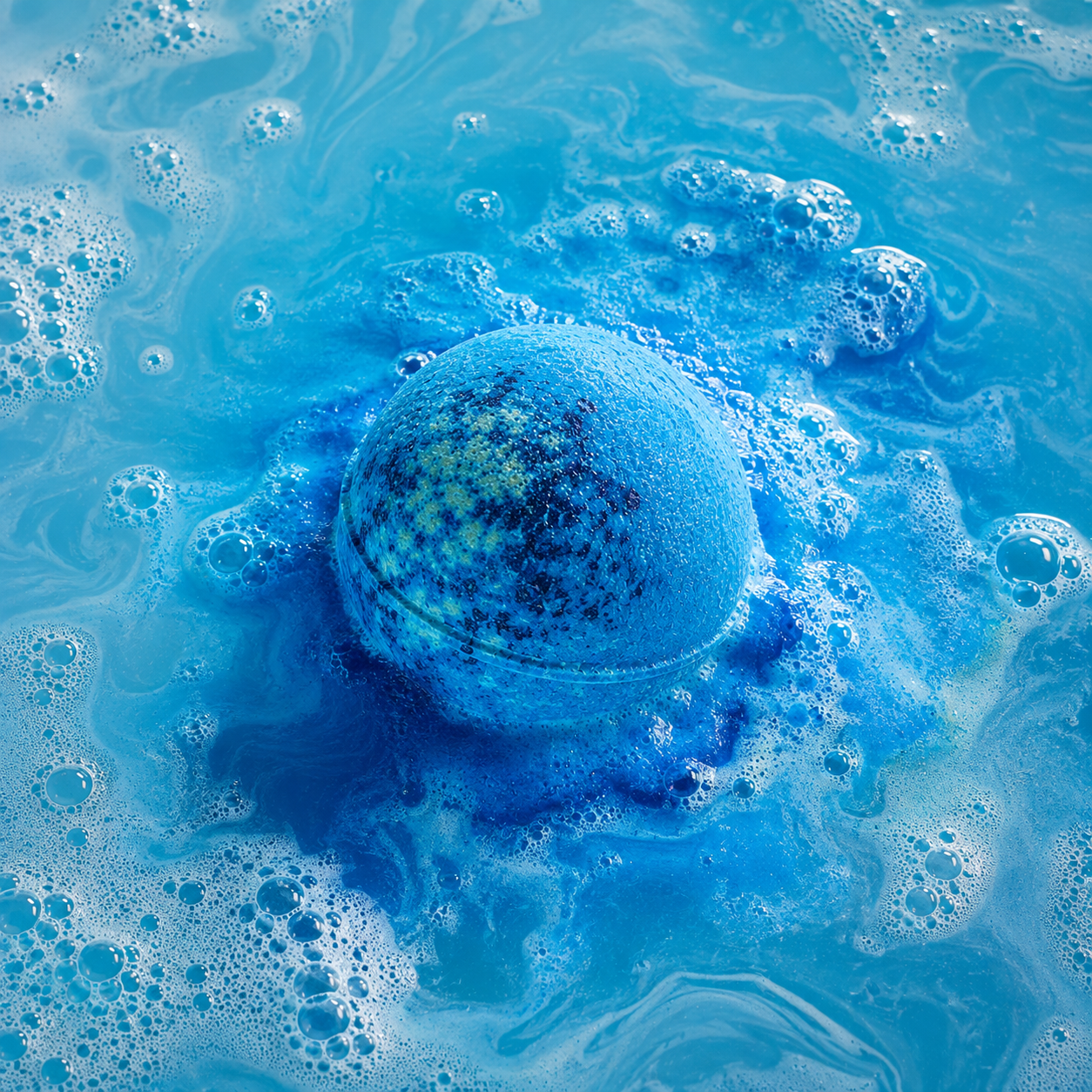 A round bath bomb or bath salt sphere floating in bright blue foamy water, with a mottled blue and yellow surface and dissolved pigment around it.