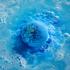 A round bath bomb or bath salt sphere floating in bright blue foamy water, with a mottled blue and yellow surface and dissolved pigment around it.