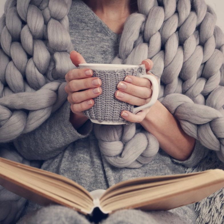 A person wrapped in a grey knit blanket, holding a cup and reading an open book.