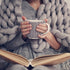 A person wrapped in a grey knit blanket, holding a cup and reading an open book.