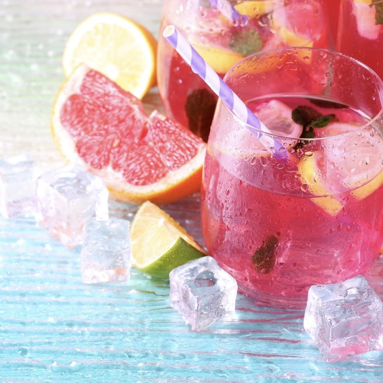 A glass of pink lemonade with ice cubes and lemon slices, alongside a purple straw and additional lemon and grapefruit segments in the background.