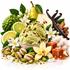 A styled food product shot featuring green pistachio ice cream scoops in a bowl, surrounded by green pistachios, white flowers, lime slices, a pear, vanilla beans, cinnamon sticks, and caramel cubes on a white background.
