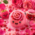 A pink rose-shaped bath bomb with a smiling face sits in a bubbly pink foam, surrounded by pink rose petals, red berries, and cute decorative bath accessories including a raspberry character and a glittery jar character.