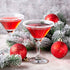 Two martini glasses filled with a red cocktail, garnished with sugar on the rim, surrounded by Christmas decorations including red baubles and artificial greenery.
