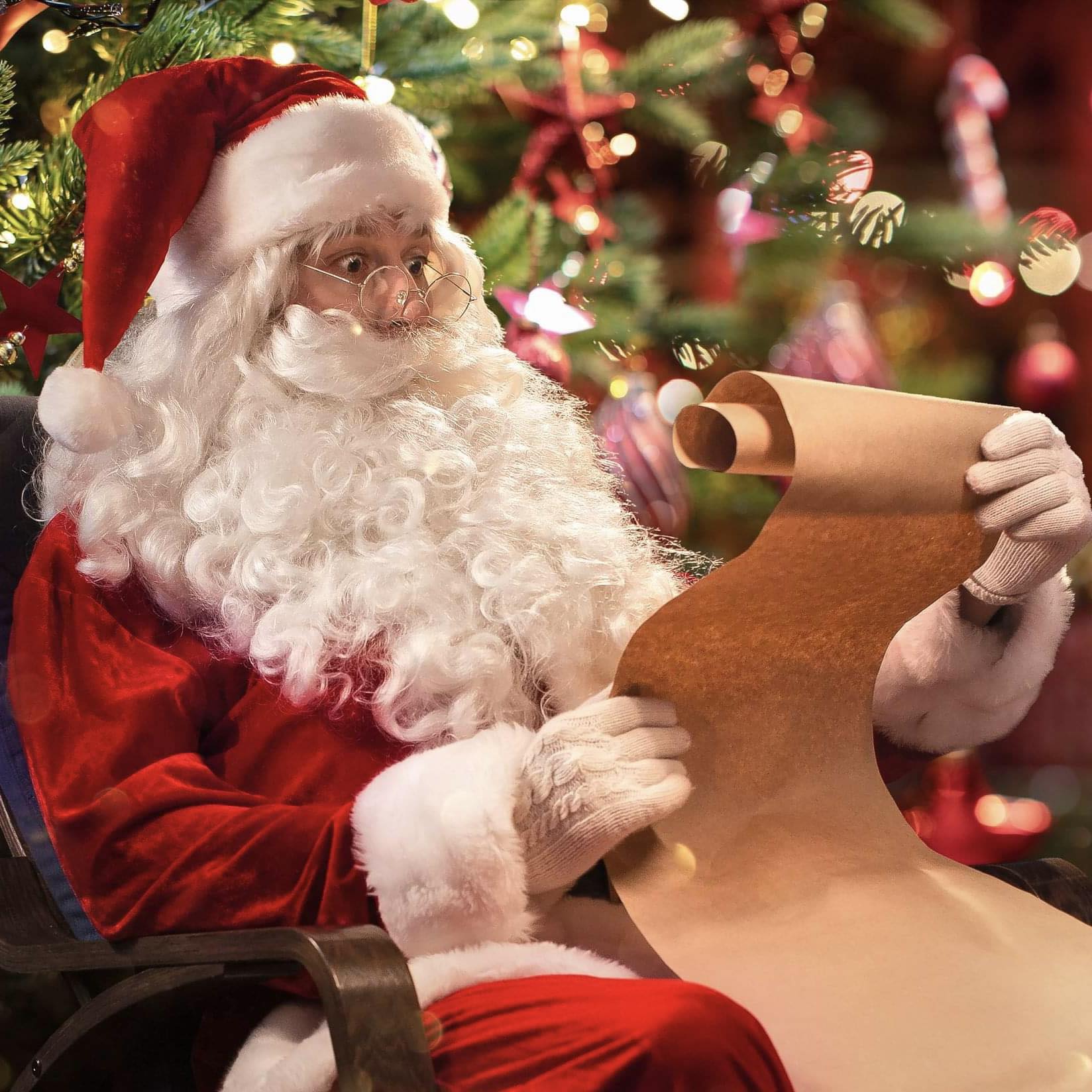 An image of Santa Claus with a white beard and red suit, sitting and reading from a scroll with a Christmas tree in the background.