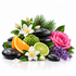 A decorative arrangement of spa-like elements on a white background: black stones, green leaves, a pink rose, white flowers, an orange slice, a lime slice, a green bumpy fruit, and a sprig of purple lavender.