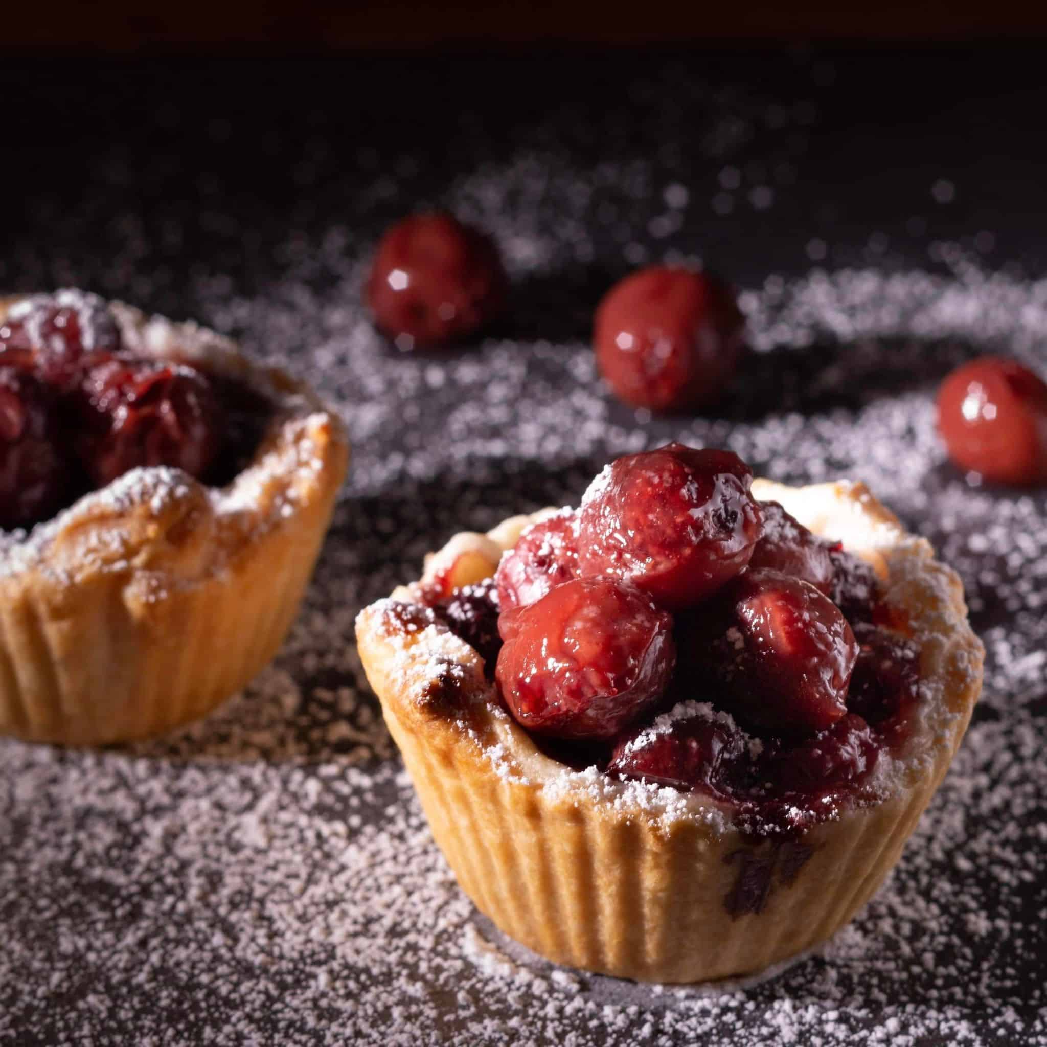 A photo depicting two small tartlet tins with a cherry and ginger frosting topping, dusted with powdered sugar, designed to represent a fragrance oil product.