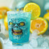 A glass with a blue liquid and ice cubes, garnished with a lemon slice, displaying a zombie face design on the glass surface.